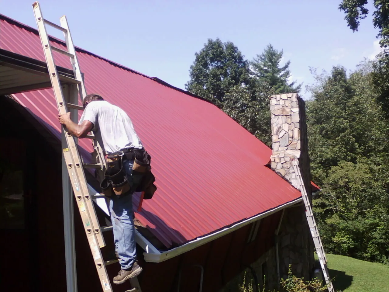 Roofer on a ladder beside a red metal roof and stone chimney — Soffit & Fascia Repair by Roofing Brothers in East Mc Keesport